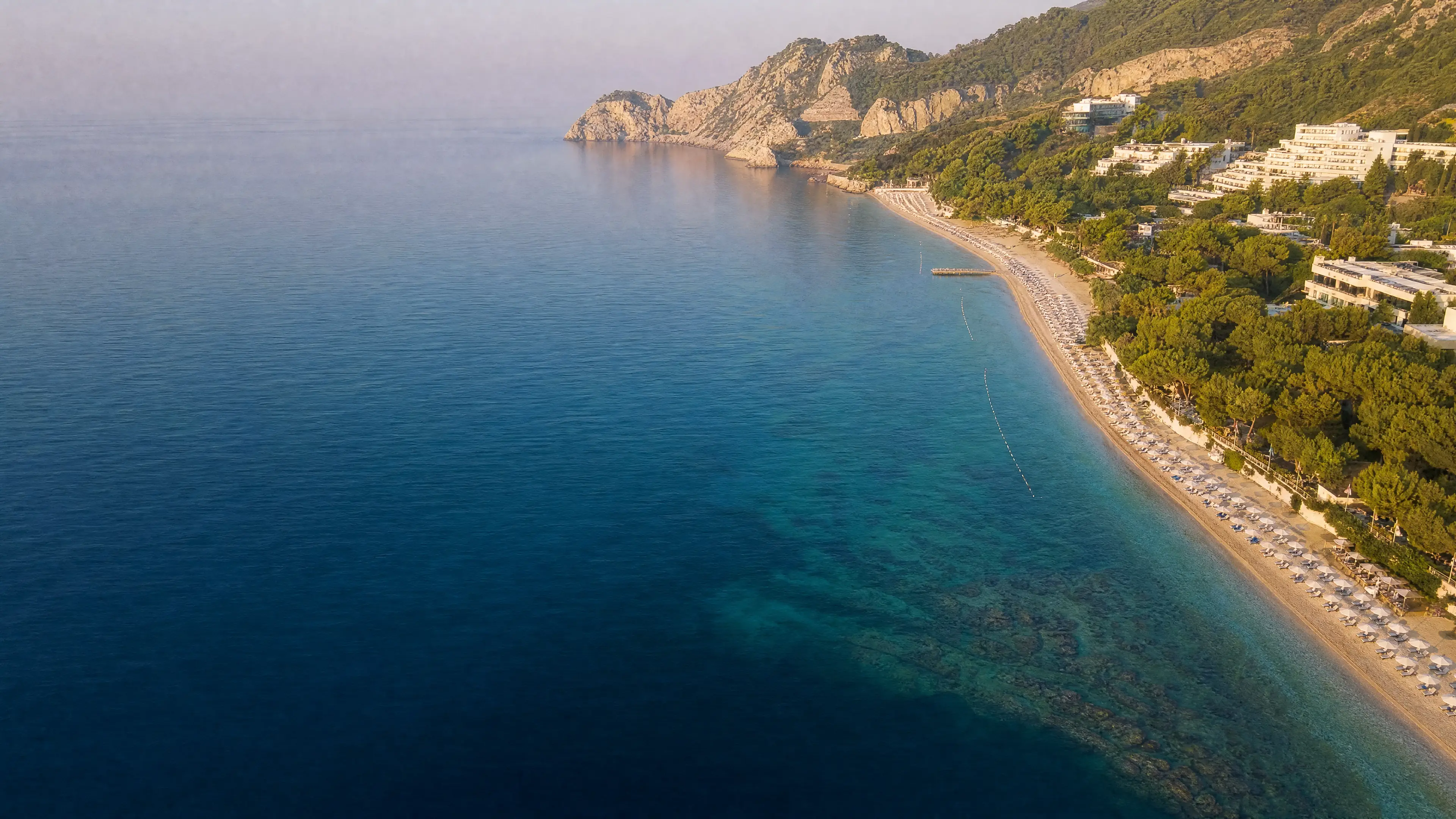 Курортное побережье с высоты: спокойное море, длинный пляж с зонтиками, отели среди зелени и скалистый мыс